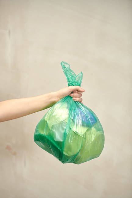A human hand holds a green plastic rubbish bag with a knot at the top against a plain, neutral background. The bag appears to contain household waste, with some visible blue and lighter green items in