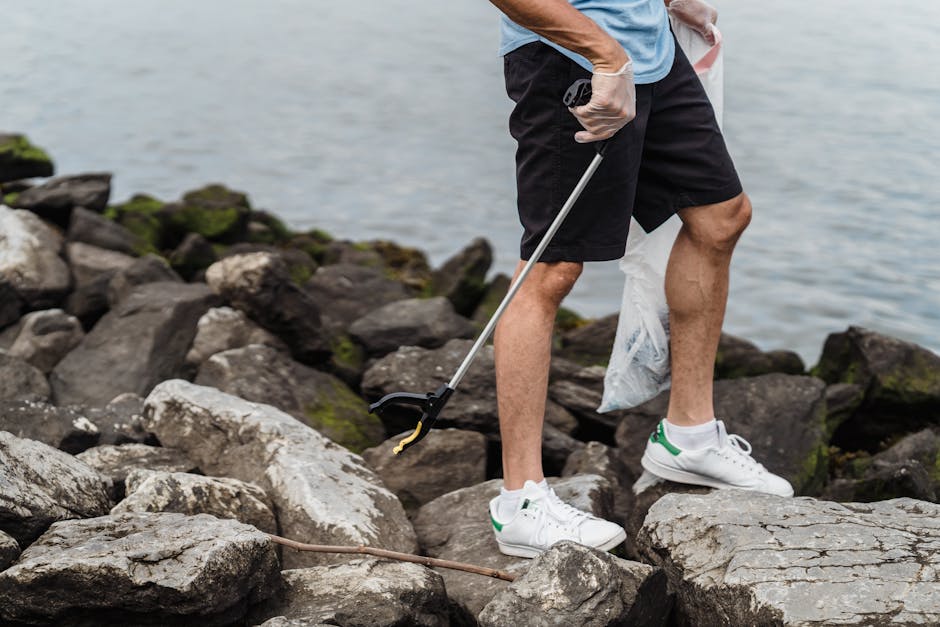 A person standing on a rocky shoreline near a body of water, wearing black shorts, a light blue T-shirt, white gloves, and white sneakers with green accents. They are holding a grabber tool in their right hand, which is extended towards a large, flat, light grey rock. In their left hand, they are holding a semi-transparent plastic bag that appears to contain waste or debris. The rocks around them are irregular in shape, with some covered in green moss or algae, and vary in shades of grey and brown. The background features calm water with a slightly rippled surface, and the scene is illuminated by natural daylight, suggesting an outdoor environment. This image depicts an individual engaged in outdoor rubbish collection or environmental cleanup along a shoreline, reflecting a focus on manual waste removal and environmental responsibility, consistent with the services of Rubbish Collection as they promote alternative waste handling methods.