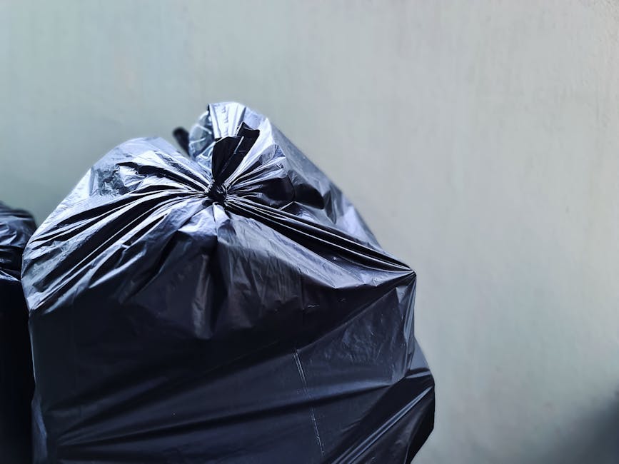 A person discarding a brown paper bag into a white plastic rubbish bin, which has a partially visible label indicating 'RUBBISH' on the side. The individual’s arm is extended, holding the bag over t